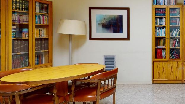 Biblioteca. La sala cuenta con paredes de tono amarillo claro, suelo de terrazo grisáceo y mobiliario de madera que incluye varias estanterías repletas de libros, dos mesas de estudio de madera clara con sillas de cuero marrón, un cuadro en la pared y un mapa de España colgado junto a una mesa auxiliar de oficina.