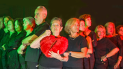 Varias personas vestidas de negro y en forma de uve miran hacia los lados. La persona del centro mira hacia el frente y sostiene un corazón roto de peluche