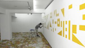 Un hombre montando una pieza en la sala de Arte Joven. Está pegando vinilos amarillos con distintas formas geométricas sobre un muro