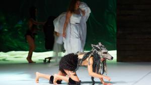 Una persona disfrazada de lobo gatea por un escenario. Detrás hay más actrices, una está vestida de blanco.