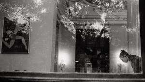 Una fotografía en blanco y negro en la que se observa una mujer detrás de una ventana de cristal