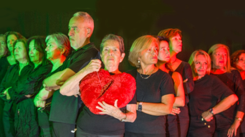 Varias personas vestidas de negro y en forma de uve miran hacia los lados. La persona del centro mira hacia el frente y sostiene un corazón roto de peluche
