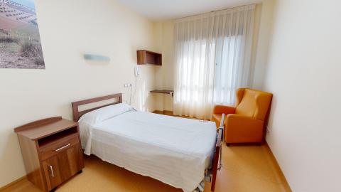 Habitación individual. Habitación individual con cama de matrimonio con ropa de cama blanca. El suelo anaranjado, muros y techo claros. Ventana al fondo con cortinas claras. Cama y mesilla de noche a la izquierda y sillón individual orejón color anaranjado a la derecha.