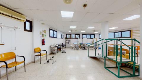 Sala de fisioterapia. Amplia sala con ventanas en lo alto. Diferentes estaciones para ejercicios de brazos, piernas, rampa, escaleras y masajes, entre otros.