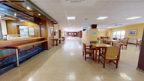 Cafetería. Cafetería. A la izquierda se encuentra la barra seguida de la cocina cerrada. A la derecha hay mesas y sillas de madera. Al fondo se alcanzan a ver unos sofás en un espacio abierto.