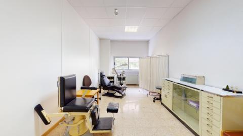 Área alargada en blanco con techos altos. Al fondo ventanas. En el muro izquierdo, dos estaciones de podología. En el muro derecho un mueble bajo largo con cajones y puertas de cristal. 