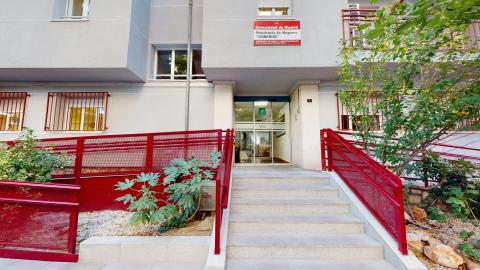 Acceso principal. Edificio gris claro. De frente escaleras, al lado izquierda rampa. Las barandillas son de metal pintadas en rojo.
