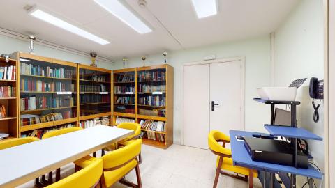 Sala pintada en verde claro con múltiples libreros de madera en el furo del fondo, al lado de la puerta, y de la izquierda. A la derecha un escritorio azul con ordenador e impresora. Al centro una mesa rectangular blanca con sillas de madera tapizadas en amarillo.