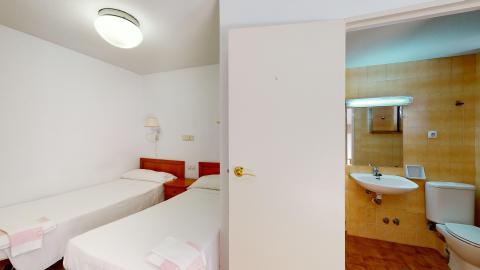 Habitación doble en blanco. A la izquierda dos camas individuales con una mesilla de noche al centro. A la derecha se ve el acceso al aseo con la puerta abierta. De frente el lavabo con espejo y a la derecha el inodoro.