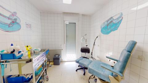Sala en mosaicos blancos con mobiliario blanco y azul. Al fondo una ventana angosta de suelo a techo. A la izquierda un muebla con los instrumentos. A la derecha la silla de podología en azul claro. 