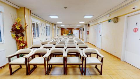 Es un espacio amplio rectangular. Se puede dividir con puertas plegables hacia la mitad de la habitación. Al fondo se ve el muro y el altar en madera de la capilla. Se ven múltiples filas de cinco sillas alineadas en montaje auditorio. A la derecha puertas hacia servicios diferenciados de señores y señoras. 