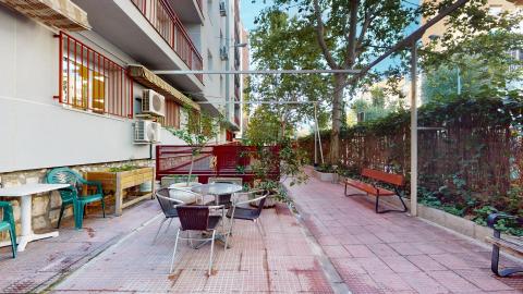 Espacio alargado de terraza con suelo rojo. A la derecha división con plantas y árboles. A la izquierda el edificio de la residencia con los muros en gris claro y herrería en rojo. Hay bancas de madera y mesas y sillas, macetas y zona de huerto. Hay posibilidad de cerrar con toldo para dar sombra. 