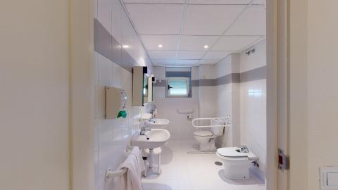 Baño y aseo. Espacio alargado en azulejos y mobiliario blanco. En el muro izquierdo hay dos lavabos con espejos con almacenamiento en el muro. A la derecha el bidet y el inodoro. Al fondo la ducha y una pequeña ventana.