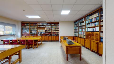 Biblioteca. Amplia biblioteca. A la izquierda ventanales que dan al exterior. Al fondo y a la derecha libreros de suelo a techo en madera. Al centro mesas y sillas de trabajo de madera. En una de las mesas hay una máquina de escribir.