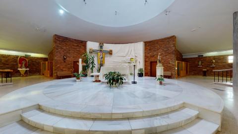 Capilla Amplia capilla circular. Los muros son de ladrillo caravista El techo es alto y blanco con una cúpula sobre el altar. El suelo es claro y hay una amplia plataforma circular de mármol blanco en el centro, debajo del altar. Hay un gran crucifijo al fondo, varias imágenes religiosas y macetas con plantas frondosas.