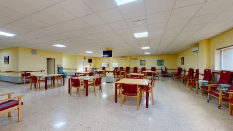 Centro de día. Amplia sala de estar en color amarillo claro. Bien ventilado e iluminado. Hay varias sillas, mesas y butacones distribuidos en distintos acomodos.