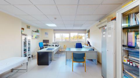 Consulta médica. Amplia sala con dos escritorios de atención. Ventanas exteriores a lo lardo del muro del fondo. Archiveros, armarios y libreros en distintos espacios y una camilla de exploración a la izquierda.