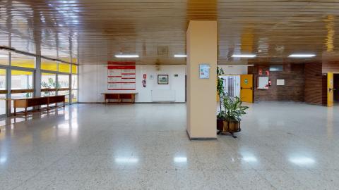 Acceso y recepción. Espacioso acceso y recepción. A la izquierda las puertas de cristal que dan al exterior. Al fondo un muro blanco con puertas y el directorio de la residencia. Al centro una columna y macetas. El techo es de madera.