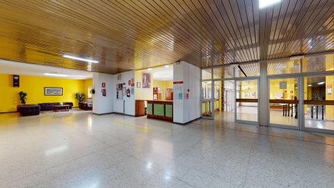Acceso y recepción. Vista complementaria de la recepción. De frente están las puertas de acceso de cristal. A su izquierda el mostrador de atención seguido de un salón de espera con muro amarillo y sofás negros de piel sintética.