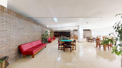 Sala de estar. Amplia sala con techos altos blancos, suelos claros y muros de ladrillo caravista. Hay sofás de piel sintética roja a lo largo del muro izquierdo. Al centro hay varias mesas de cuatro para juegos. Hasta la derecha es todo ventanal para dar luz y ventilación. Se aprecian varias macetas con plantas altas y frondosas..