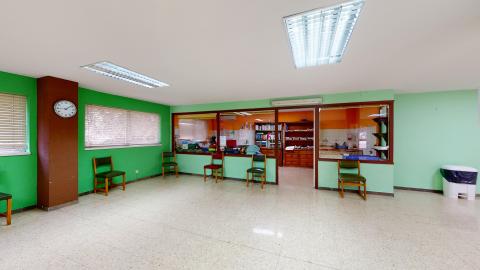 Terapia ocupacional. Sala en verde diáfana con sillas acomodoadas alrededor. Hay un espacio cerrado con medio muro y ventanales para el almacenaje de material y zona de trabajo. A la izquierda ventanas que dan al exterior.