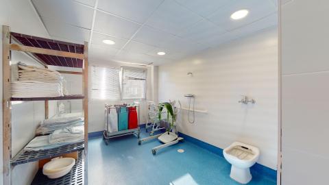 Aseo y baño geriátrico. Amplio baño con suelo azul y muros y paredes en blanco. Hay una ventana grande que da luminosidad. Es completamente accesible.