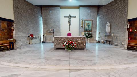 Capilla. Vista del altar de la capilla. Muros de ladrillo caravista gris claro. Techos muy altos. Al centro un gran crucifijo en el centro. Delante el altar con grabado metálico. Muy luminoso. Hay varias imágenes religiosas en cuadro y esculturas pequeñas y medianas..