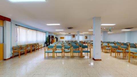 Sala de actividades. Amplia sala en azul claro con la mitad de los muros con amplias ventanas con cortinas blancas traslúcidas. Hay sillas de madera tapizadas en verde acomodadas en auditorio.