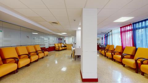 Sala de estar. Amplia sala luminosa. Al centro hay columnas. A la izquierda un muro de cristal opaco con butacones individuales amarillos acomodados en fila. A la derecha ventanas de piso a techo con cortinas traslúcidas en azulo, rojo y blanco. En frente, más butacones en fila.