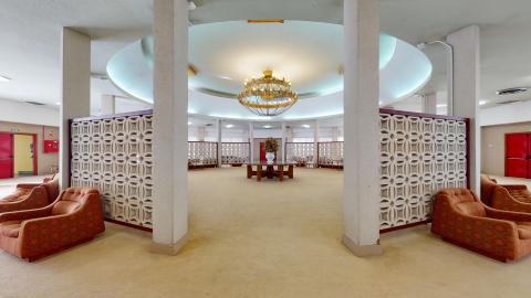 Salón. Amplio salón circular delimitado por una celosía de hormigón en blanco. Fuera de la celosía hay sillones individuales largos tapizados en anaranjado. Al centro, una gran mesa circular de madera debajo de una gran lámpara de araña. El suelo es beige.
