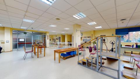Fisioterapia. Amplio espacio con muros en amarillo claro. Hay ventanas grandes y luminosas. La puerta es de cristal opaco dando luz pero intimidad. Hay múltiples estaciones para ejercitar diferentes músculos y movimientos.