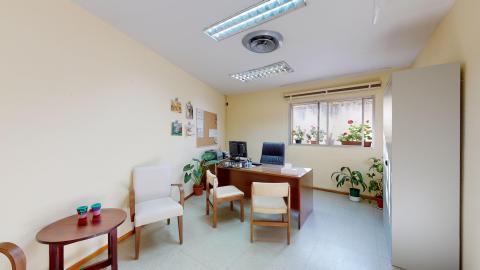 Consulta de psicología. Sala luminosa en colores claros y madera. Al fondo ay una ventana luminosa con macetas con flores en el bordillo de la ventana y debajo de la misma. El escritorio es de madera. En el muro izquierdo hay una mesa con dos sillas.