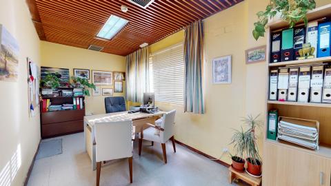 Trabajo social. Sala acojedora en colores beige y techo de madera. A la derecha una amplia ventana muy luminosa con cortinas. Al fondo un librero en marrón oscuro, rodeado de cuadros. Al centro el escritorio con sillas en blanco y madera. Frente a la entrada, un librero con archivos. Hay múltiples cuadros y macetas.