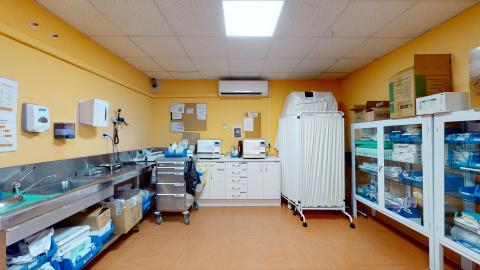 Consulta enfermería. Espacio pintado de amarillo. A la izquierda una barra de acero inoxidable con lavabo. Al fondo un mueble de cajones en blanco y biombos. A la derecha, muebles de cristal con puertas para material sanitario.