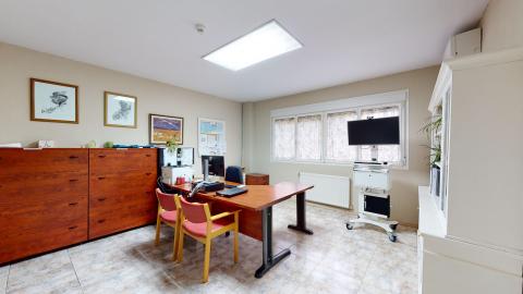 Consulta médica. A la izquierda dos muebles de madera rojiza con cajones grandes y cuadros en el muro. Al fondo, una ventana amplia y luminosa. A la derecha un mueble librero y cajonera en blanco. Al centro el escritorio. Los muros en beige claro.