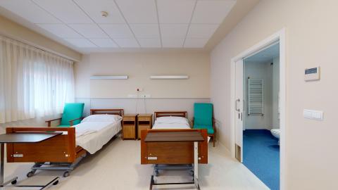 Habitación doble. A la izquierda una gran ventana luminosa con cortinas blancas. Al centro, dos camas individuales con mesillas de noche al centro y butacones individuales en verde a los extremos. A la derecha, tras una amplia puerta corrediza, el baño.