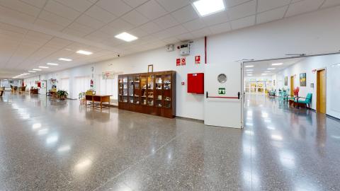 Acceso y recepción. Vista complementaria de los espacios que unen las distintas estancias. Pasillos muy amplios y luminosos. Sillas distribuidas a lo largo y un gran mueble de madera que exhibe trabajos de los residentes.