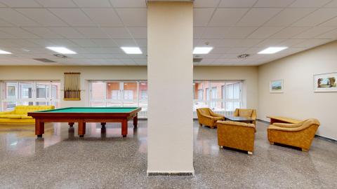 Sala polivalente. Amplia sala con techos altos blancos, suelos claros y muros de ladrillo caravista. Hay sofás de piel sintética roja a lo largo del muro izquierdo. Al centro hay varias mesas de cuatro para juegos. Hasta la derecha es todo ventanal para dar luz y ventilación. Se aprecian varias macetas con plantas altas y frondosas..