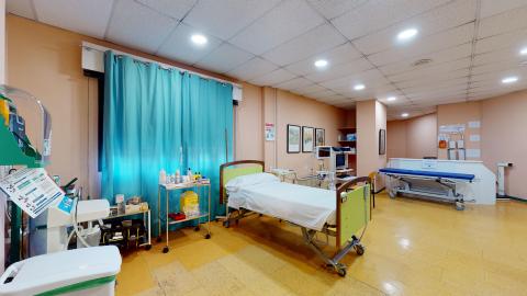 Consulta de enfermería. Espacio amplio con suelos en amarillo, muros en marrón claro y techos blancos. Hay una cama y una camilla, instrumental sanitario disponible. Una ventana grande luminosa y cuadros en uno de los muros.