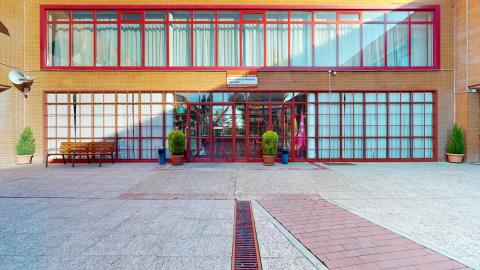 Acceso. Fachada de acceso. Se ven dos plantas de un edificio de ladrillo caravista. La planta baja tiene todo el muro de puertas de cristal con herrería en rojo. La primera planta tiene ventanales de piso a techo también con herrería en rojo. En frente hay una banca de madera y cuatro macetas grandes.