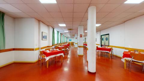 Comedor. Comedor alargado con muros en blanco, suelo en anaranjado y detalles en madera. Hay columnas que dividen el espacio a lo largo por la mitad. En el muro izquierdo hay múltiples ventanas con cortinas blancas y verdes. Las mesas son para tres y cuatro comensales y tienen manteles en blanco y ojo. A la derecha están las puertas de la cocina.
