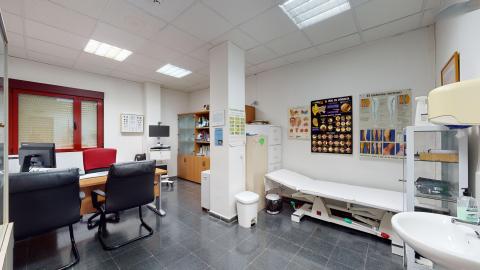 Consulta médica. Espacio rectangular en blanco. A la derecha hay un lavabo y un armario de cristal. De frente se ubica la camilla seguida de archiveros de metal y libreros de madera. A la izquierda hay una ventana amplia con marco rojo y, delante, el escritorio. En los muros hay paneles informativos del sistema venoso, del oído, de la garganta y un panel de optometría..