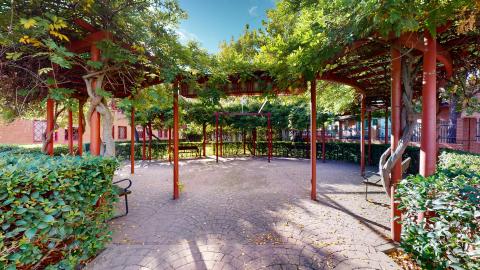 Jardín. Patio circular delimitado por arbustos al centro de construcciones de ladrillo caravista.. Al centro, el suelo está adoquinado. Hay tres bancas de madera alrededor. Hay una estructura metálica pintada en cojo con enredaderas que dan sombra.