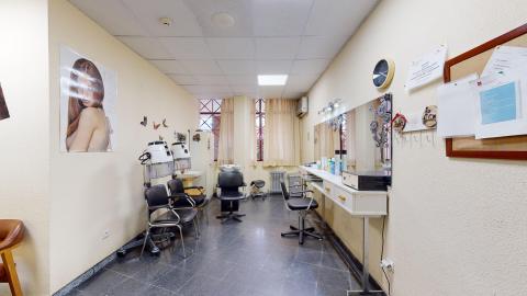 Peluquería. Espacio alargado pintado en beige. Al fondo hay ventanas con cortinas beige. En frente de ella, una estación de lavado. A la izquierda, dos estaciones de lavado seguidas de un lavabo y un armario blanco. A la derecha tres estaciones de corte frente a un gran espejo. En los muros, un poster y una pizarra de corcho.