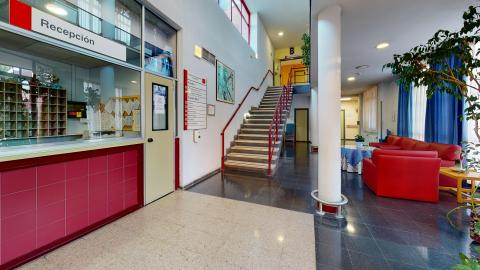 Recepción. Estancia de acceso y distribuidora de espacios decorada en blanco, rojo y azul. A la izquierda la recepción, de frente las escaleras, a la derecha un salón de piel sintética roja y ventanas exteriores.