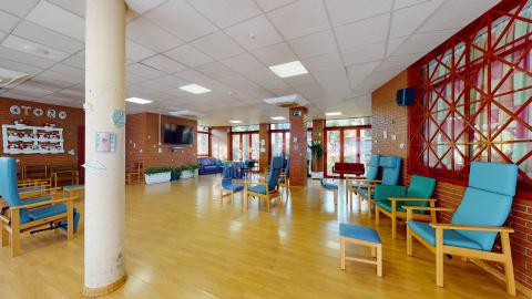 Sala de estar. Amplia sala diáfana con muros en ladrillo caravista, techos blancos y suelo de tarima. Tiene amplias ventanas exteriores con marcos en rojo. En uno de los muros cuelga un televisor grande. Distribuidos alrededor, butacones, sillas, reposapiés y sofás en piel sintética azul con madera.