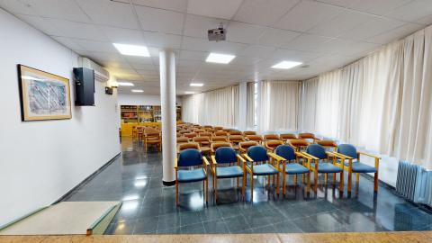 Salón de actos. Amplia sala diáfana en blanco. Al fondo, hay libreros de madera. A la derecha todo son ventanas exteriores con cortinas blancas. Al centro, sillas acomodadas en auditorio y una columna cilíndrica blanca. La foto se ha hecho desde el escenario qe tiene una tarima de madera y es accesible por una rampa.