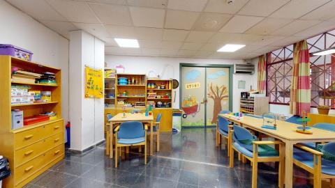 Terapia ocupacional. Sala blanca con mobiliario de madera clara. En los muros izquiero y al fondo hay libreros con material. Al fondo una puerta de acceso con decoración de otoño. A la derecha amplias ventanas exteriores. Al centro, dos mesas, una con cuatro sillas y otra con seis.