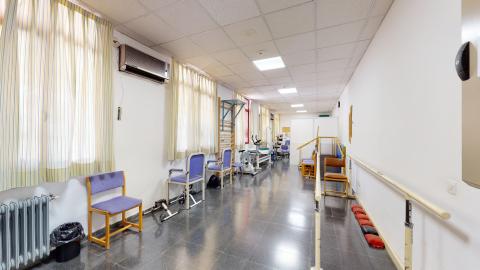Fisioterapia. Sala alargada pintada en blanco. En el muro izquierdo hay múltiples ventanas exteriores grandes con cortinas claras. Debajo de estas ventanas, hay estaciones de fortalecimiento. A la derecha soporte para practicar la marcha.