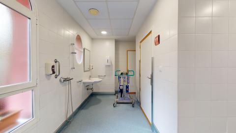 Baño. Se aprecia un baño con muros en azulejos blancos, techo blanco y suelo azul. En el muro izquierdo hay una ventana grande luminosa de cristal opaco y otra ventana circular más adelante al ladeo del espejo del lavabo. Entre ambas ventanas está la ducha completamente accesible. Se aprecian dos puertas blancas al lado derecho, una para inodoro de señoras y otra de acceso.